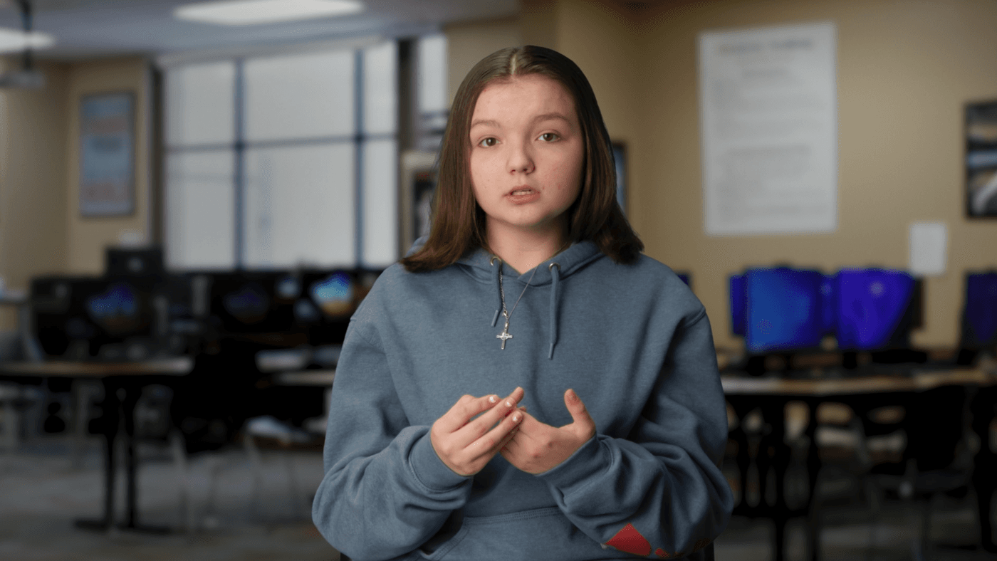 A young girl with long brown hair, wearing a blue hoodie and a cross necklace, is speaking in a blurred classroom setting with computer monitors in the background.