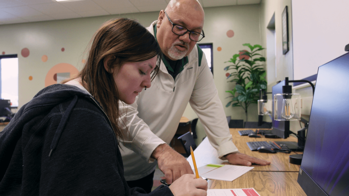 A middle-aged man with a white beard and glasses leans over a desk, pointing to a document that a young woman with long brown hair is writing on in an office setting.