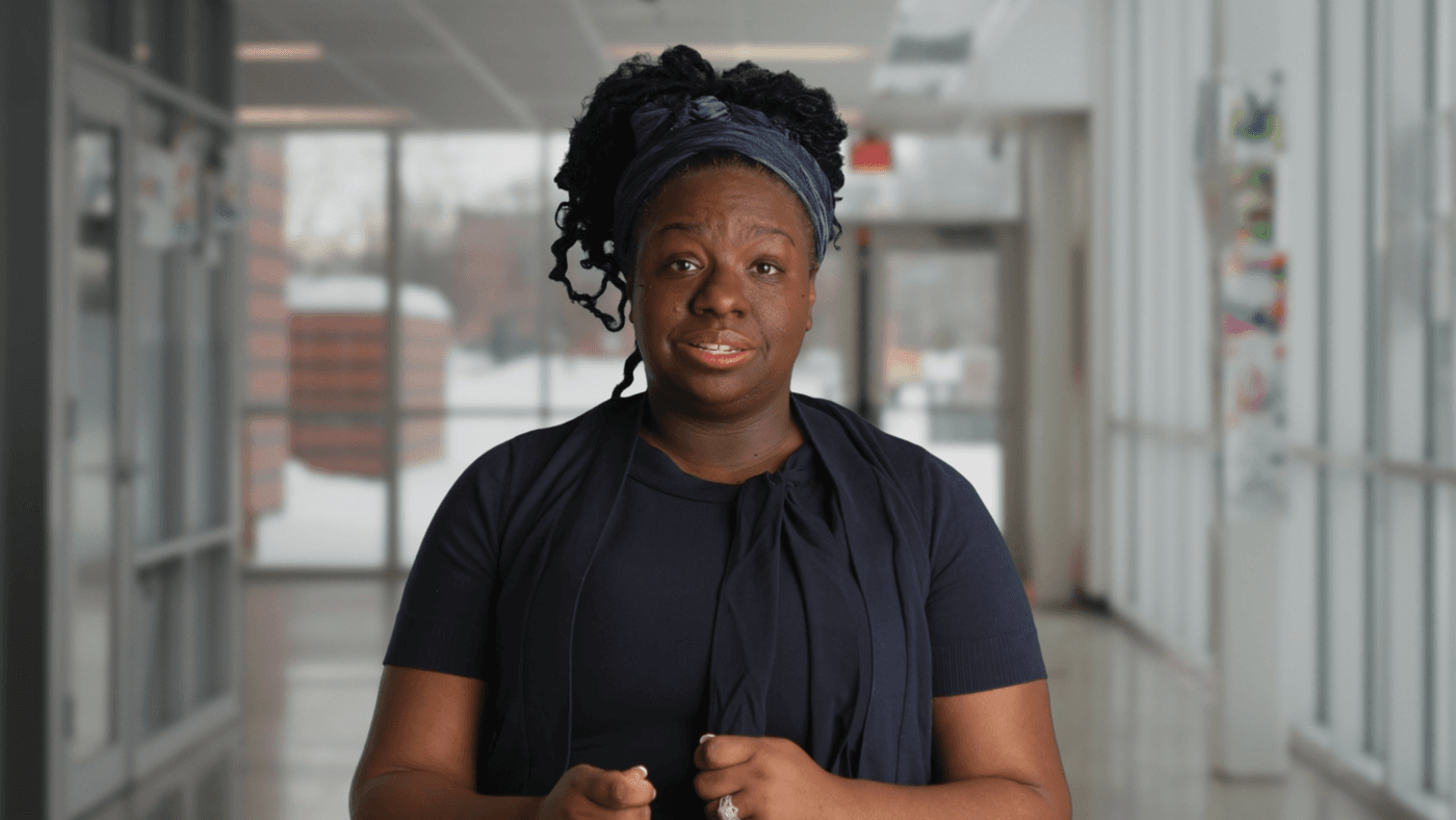 A medium shot of a Black woman with a blue headscarf and a navy blue outfit speaking while standing in a brightly lit corridor with floor-to-ceiling windows showing snow outside.