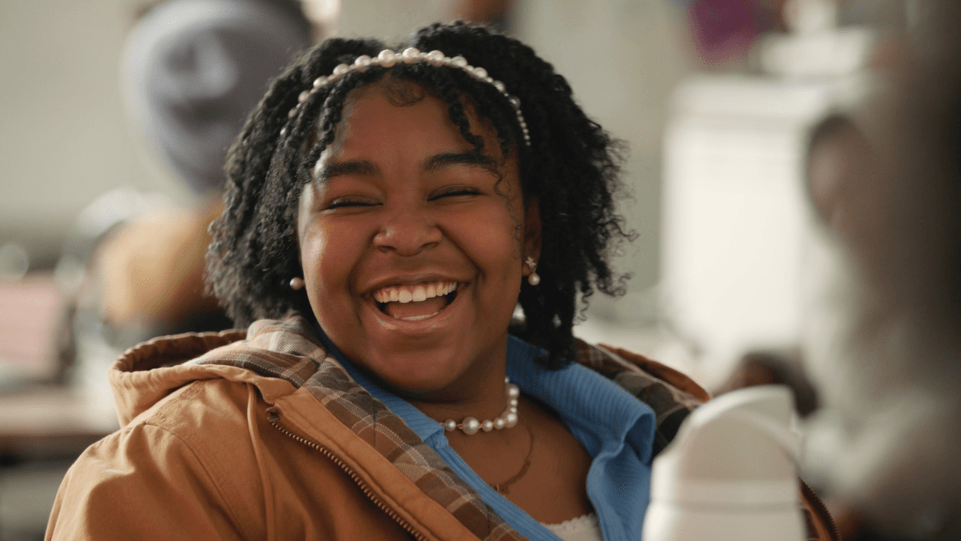 A young Black woman with dark, textured hair adorned with a pearl headband laughs joyfully while wearing a blue collared shirt under a tan jacket and a matching pearl necklace.