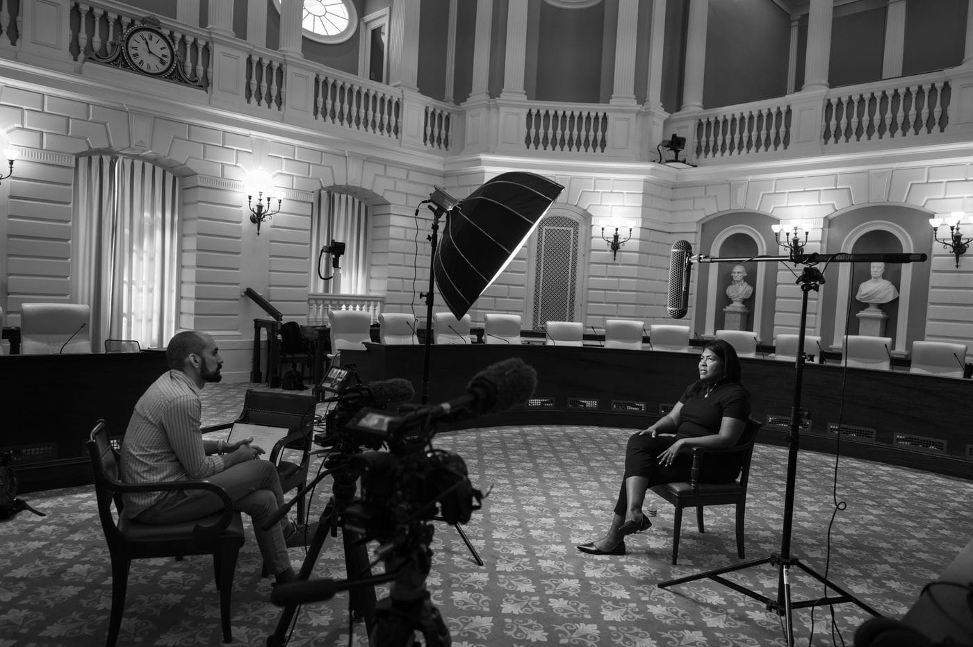 A person interviews a woman seated in a chair in a grand, circular hall with neoclassical architecture, captured by camera and lighting equipment.