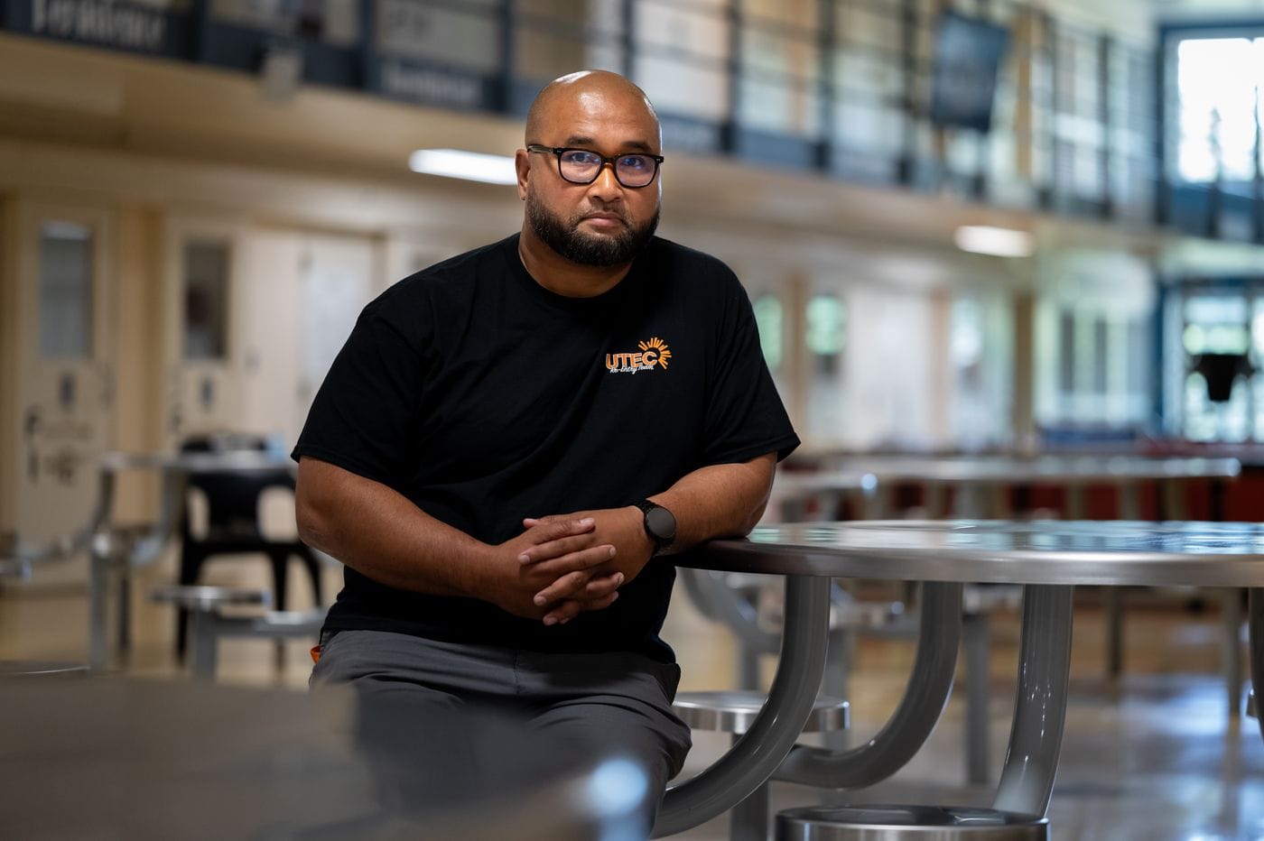 A bald man with a beard and glasses wears a black t-shirt with a UTEC logo and sits with his hands clasped at a metal table in a large, bright open space.