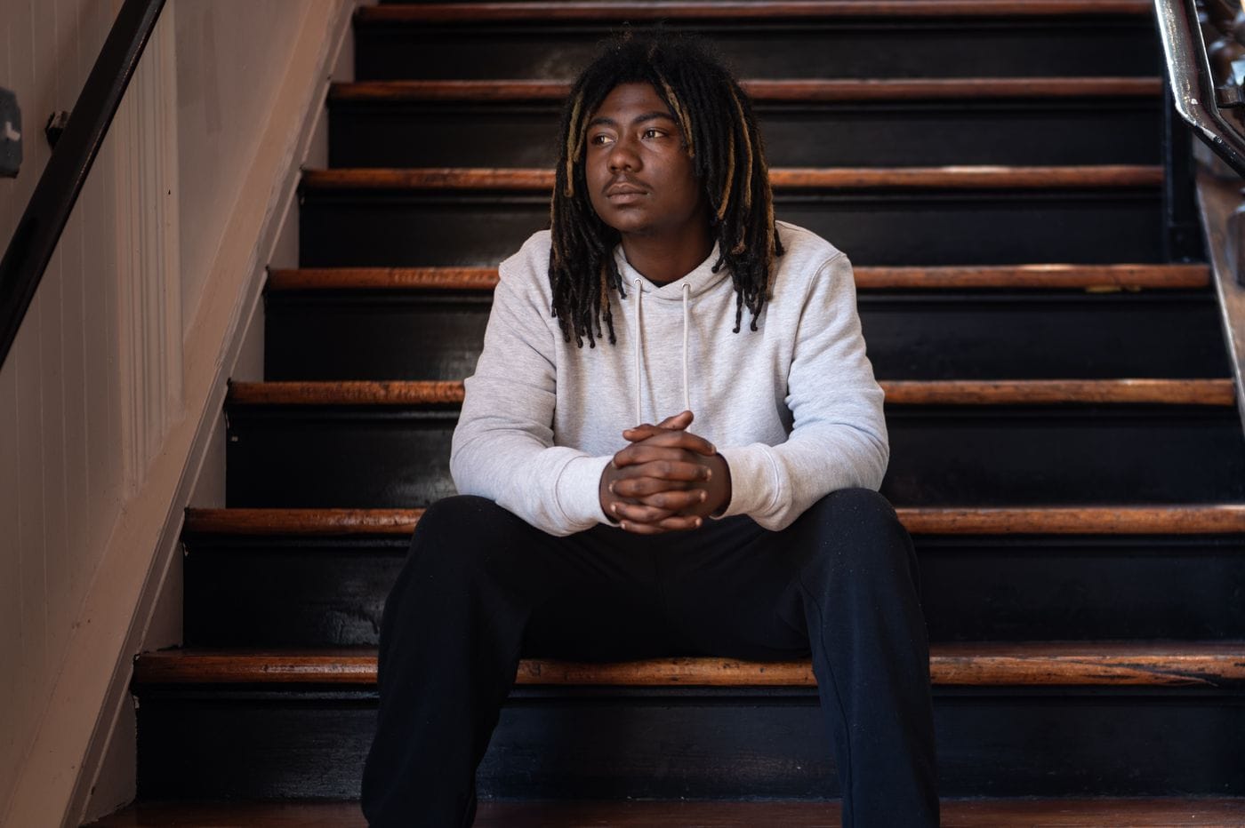 A young Black man with dreadlocks wearing a light grey hoodie and black pants sits with his hands clasped on a set of dark wooden stairs.