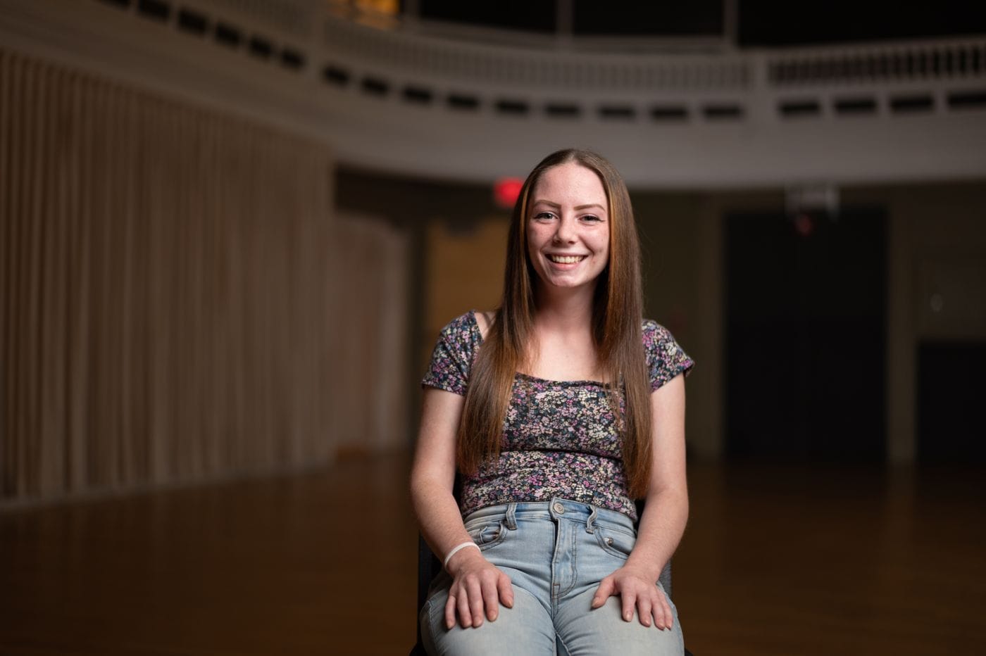 A young woman with long, straight brown hair and freckles smiles while sitting in a chair in a dimly lit, spacious hall with a balcony and draped curtains in the background.