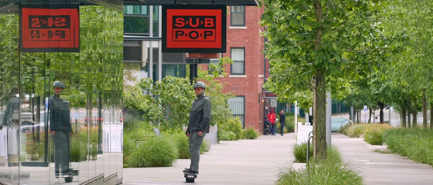 A person in a gray jacket and helmet rides an Onewheel on a sidewalk past a Sub Pop store sign, with its reflection visible in a glass wall on the left.