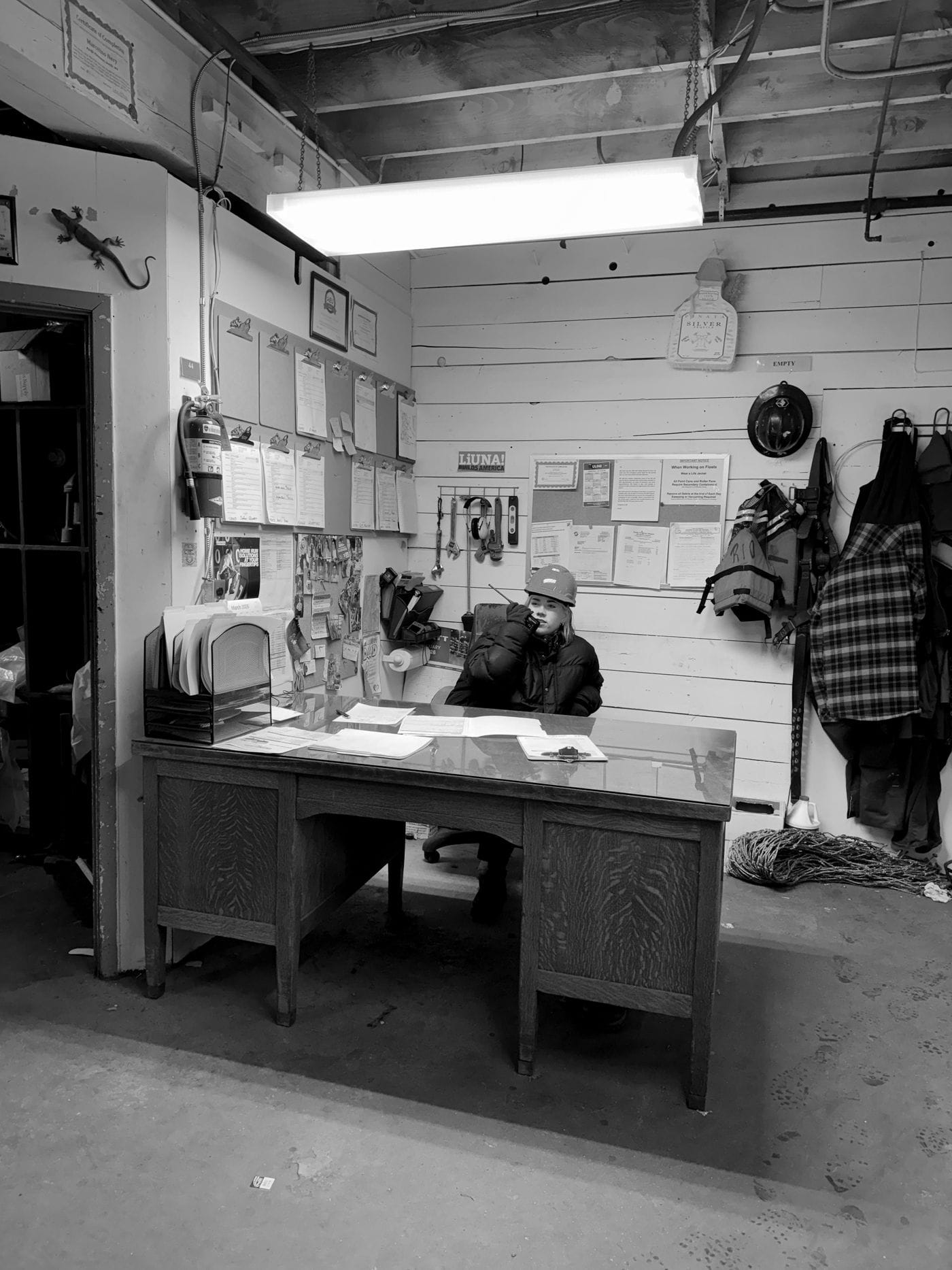 A woman wearing a hard hat and dark jacket sits at an old wooden desk in a rustic office, speaking into a two-way radio while surrounded by paperwork, safety gear, and construction-related posters.